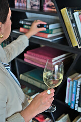 A woman selecting a book from a shelf while holding a glass of white wine, ideal for lifestyle and leisure promotions with copy space.