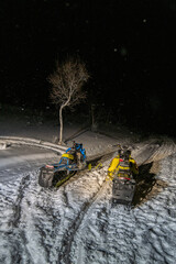 Two bright snowmobiles stand in the light of headlights illuminating the night slope and a lonely tree growing on it