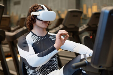 Portrait of young man working out with exercise bike in gym and wearing VR headset gesturing and...
