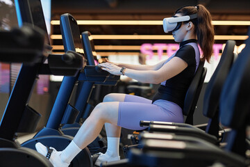 Full length side view portrait of futuristic workout on exercise bicycle with girl wearing VR...