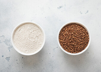 Ceramic bowls with dry raw brown buckwheat seeds and flour powder on kitchen table.Macro.