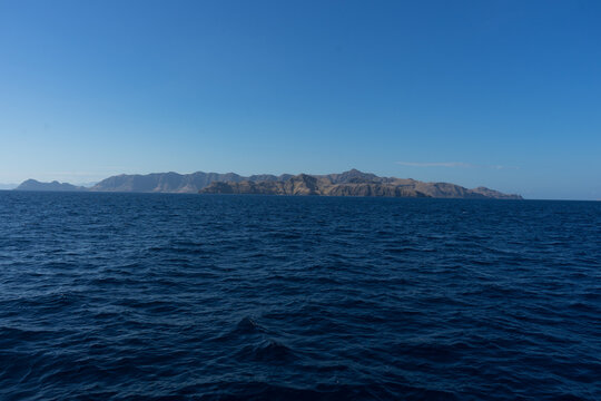 A serene view of a vast ocean with a rugged island in the distance under a clear blue sky.