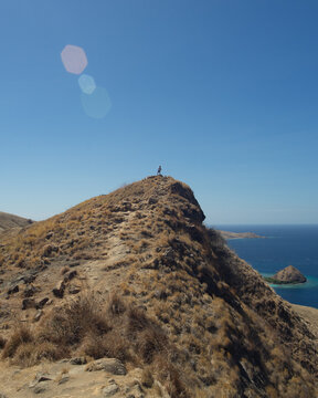 A lone person stands on the peak of a dry, grassy hill overlooking the sea.