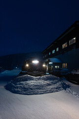 A snowcat, waiting for a group of skiers and snowboarders, clears the snow near the entrance to the hotel early in the morning during a snowfall and illuminates everything with headlights