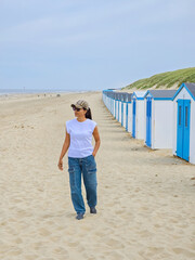A stylish woman strolls gracefully, passing by a vibrant row of beach huts lining the Texel,...