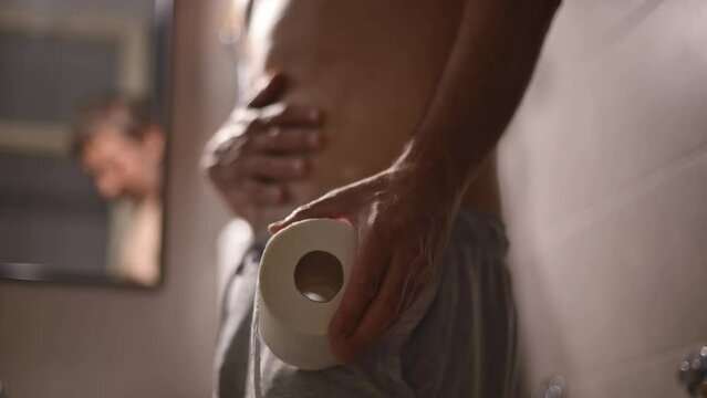 Constipation concept, constipated mature adult man standing in bathroom with roll of toilet paper