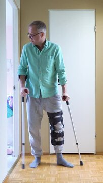 Middle-aged man with leg injury walking on crutches at home. Man wearing leg brace knee support adjustable strap fracture fixator