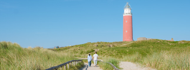 Two people walk down a path next to the lighthouse in Texel, Netherlands, with waves crashing...