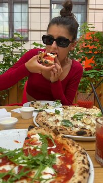 A beautiful young woman wearing sunglasses enjoys pizza on the restaurant terrace.