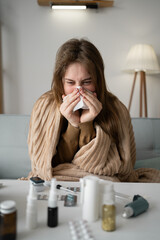 Wrapped in a blanket, a woman sits on the couch at home, fighting a seasonal cold. With a tissue in...