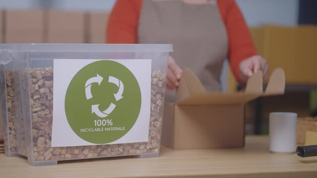 The focus is on a "100% Recyclable Materials" label, showcasing a small business's sustainable practices, A woman in the background uses eco-friendly packaging, water hyacinth cushioning