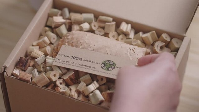 A woman places a "100% Recyclable Materials" label on a cardboard box filled with eco-friendly water hyacinth cushioning, demonstrating sustainable and environmentally friendly business practices