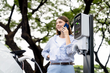 Young woman talking on smartphone while recharging electric car battery charging from EV charging...