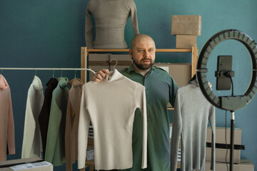 The store owner broadcasts online and talks about the quality of the clothes, holding sweaters in...