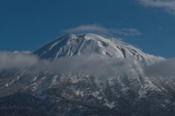 Snow-capped peak of Yotei volcano rising from the clouds and illuminated by the bright morning sun against a blue background in Niseko, Japan