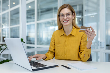 Smiling woman in glasses using a laptop and holding a credit card while sitting at an office desk....