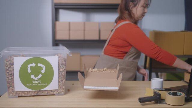 The focus is on a label stating "100% Recyclable Materials" highlighting the sustainable practices of a small business, In the background, a woman packs a product in a cardboard box with eco-friendly