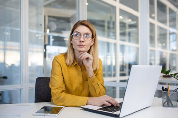 Thoughtful businesswoman wearing glasses and yellow shirt working on a laptop in a modern office...