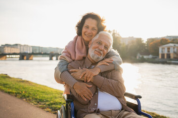 Old man in wheelchair walking with caregiver senior woman on road in park. Elderly family couple...