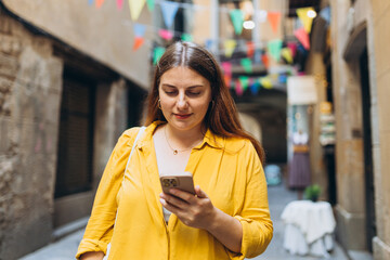 30s woman tourist dressed in casual look holding smartphone gadget in hand for communicate on the city street. Happy woman chatting on phone and relaxing outdoors. Urban, people concept. Old town