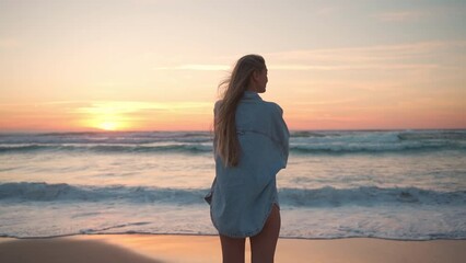 Carefree woman standing on seashore at windy beach during sunset. Back view of female tourist in long denim shirt is enjoying idyllic view of seascape at sundown. 