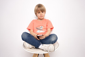 serious little boy using smartphone while sitting in a chair isolated on white background