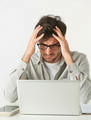 Close-up of a young man at a computer desk with a splitting headache and holding his head in his hands with an expression of pain