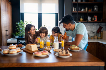 Happy parents having breakfast at kitchen table with kids at home, looking at them with love and kindness.