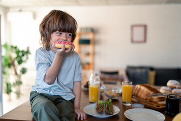 Portrait of a cute little boy eating donut during breakfast time at home. He is sitting on the table in the kitchen and looking aside.