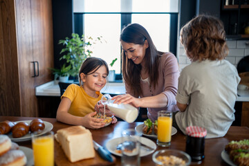 Little girl pouring milk into cornflakes cereals with mothers help. Daughter and mother preparing morning meal together. Cheerful family moments, bonding and quality time.