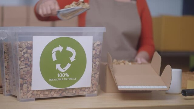 The focus is on a "100% Recyclable Materials" label, showcasing a small business's sustainable practices, A woman in the background uses eco-friendly packaging, water hyacinth cushioning