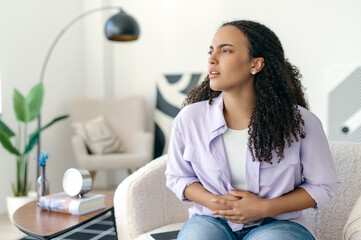 Unhappy sick young curly haired brazilian or hispanic woman in casual clothes, sitting on couch in...