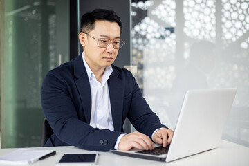 Asian mature man in a business suit working on a laptop computer in a modern office setting, focusing on his task.