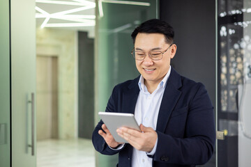 Fototapeta premium Asian mature man in a business suit smiling while using a digital tablet in a modern office setting.