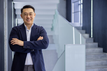 Confident asian mature man in business attire standing with arms crossed in modern office building background, showcasing success and professionalism.