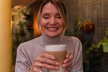 Woman with blonde hair sips cappuccino in a cafe. She is holding the glass up to her face, taking a...
