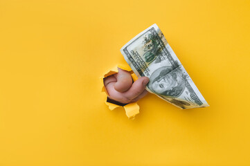 A man's hand holds dirty money through a torn hole in yellow paper. Concept of dishonest income, donation, profit and salary fraud.