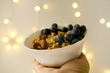 close-up female hand holds healthy food white cup, oatmeal, blueberries, nuts, almonds, peanuts,...