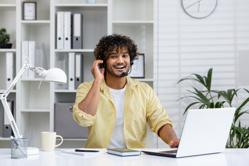 Smiling customer service representative with headset working at laptop in modern office setting,...