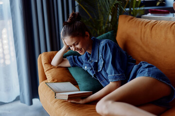 Young woman relaxing on couch with legs crossed reading an open book in cozy home environment