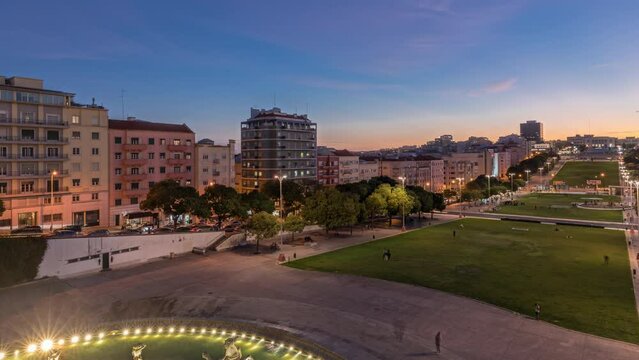 Panorama showing lawn at Alameda Dom Afonso Henriques with colorful buildings and illuminated Luminous Fountain aerial day to night transition timelapse from above after sunset in Lisbon, Portugal