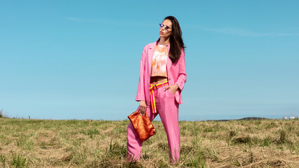 model fashion trend women. Model with pink jacket and pants suit and orange handbag with sunglasses in the sun with light blue sky background, spring summer fashion editorial