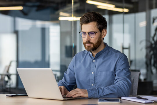 A young man in a shirt and glasses is working concentratedly in a modern office at a laptop, typing on a keyboard