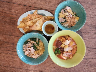 Bird's-eye view of noodles served on wooden table. Tom Yum noodles without soup and Tom Yum soup with crushed peanuts, Yen Ta Fo noodles in red soup with meatballs, fried dumplings with dipping sauce.