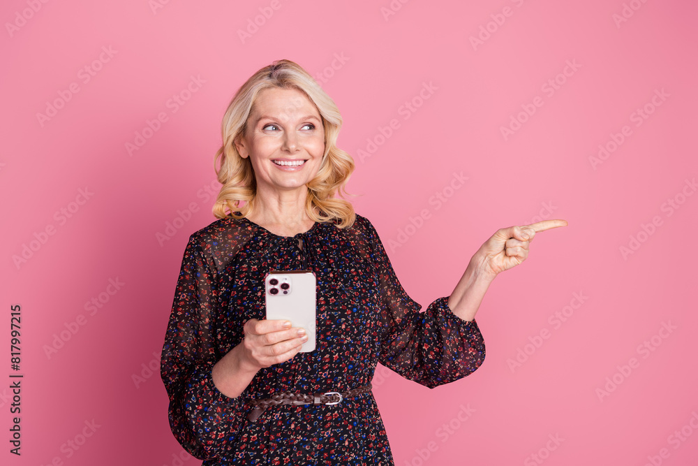 Wall mural Photo of pretty aged lady hold phone indicate finger empty space wear dress isolated on pink color background