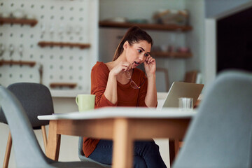 A serious young adult woman working from home on her laptop while biting on her reading glasses...