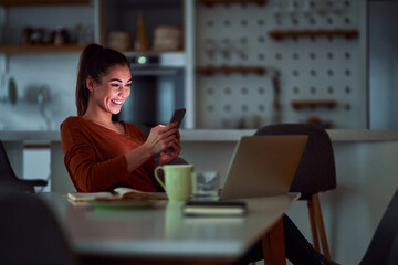 A cheerful young adult woman working late from home and using her phone