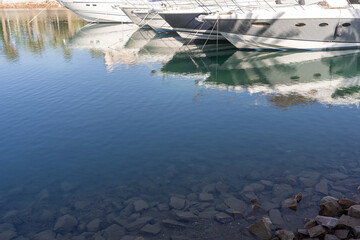 Clear water reflects luxury yachts docked in a serene marina, with rocks visible at the bottom. A peaceful scene showcasing marine leisure.