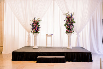 An elegant stage is set up for a wedding ceremony with two pillars with a pretty floral...