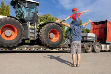 man and boy sitting on his shoulders stand in front of large tractor loaded on truck trailer. son's...